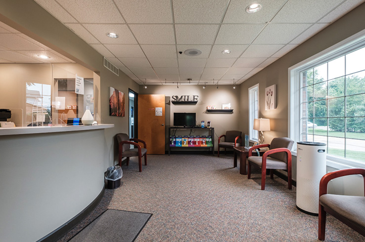 The image shows an interior view of a dental office waiting area with a reception desk, chairs, and a window.