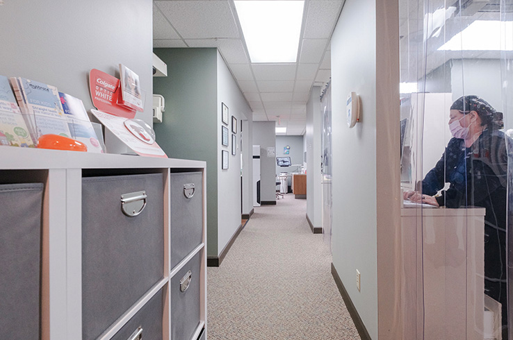 The image shows an interior hallway with a person standing at a desk, a reception area with a sign on the wall, and a door leading to another space.