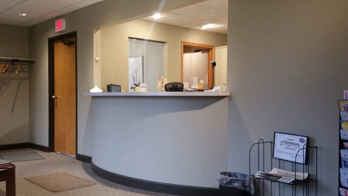 The image shows an interior space with a reception desk area featuring a countertop with a sign on top, a large wall-mounted clock above the counter, and a person seated behind the desk.