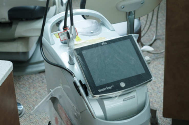 The image shows a medical device with a tablet attached to it, placed on a cart with wheels, sitting next to a dental chair.
