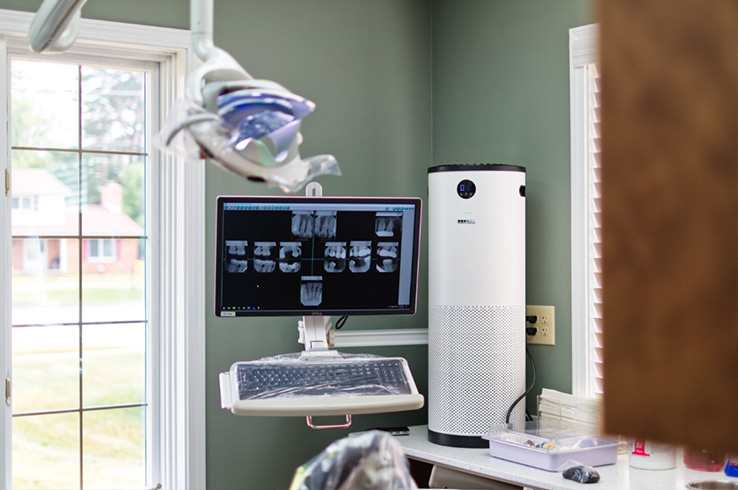 The image shows a dental office interior with a monitor displaying an X-ray image, a computer monitor on a desk, a laptop, a chair, and various dental equipment visible.