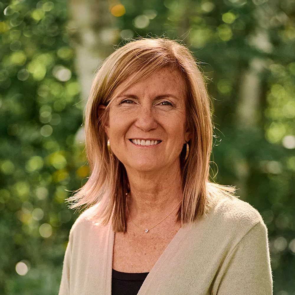 The image shows a woman with short hair wearing a light-colored cardigan and standing outdoors in front of trees, smiling slightly towards the camera.