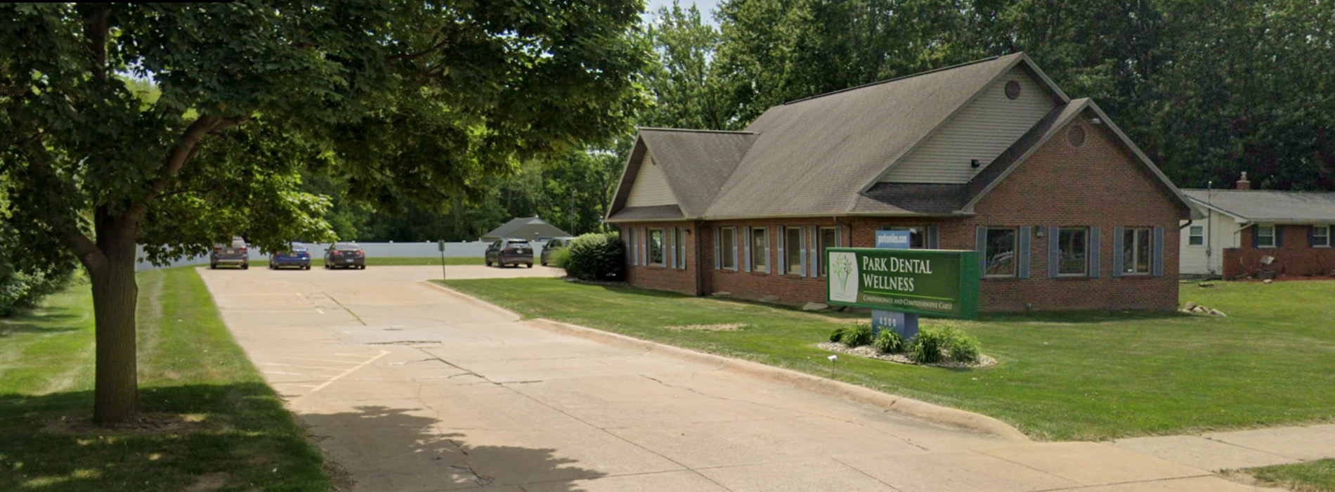 The image shows a two-story building with a covered porch, situated on a street corner with trees and a clear sky.