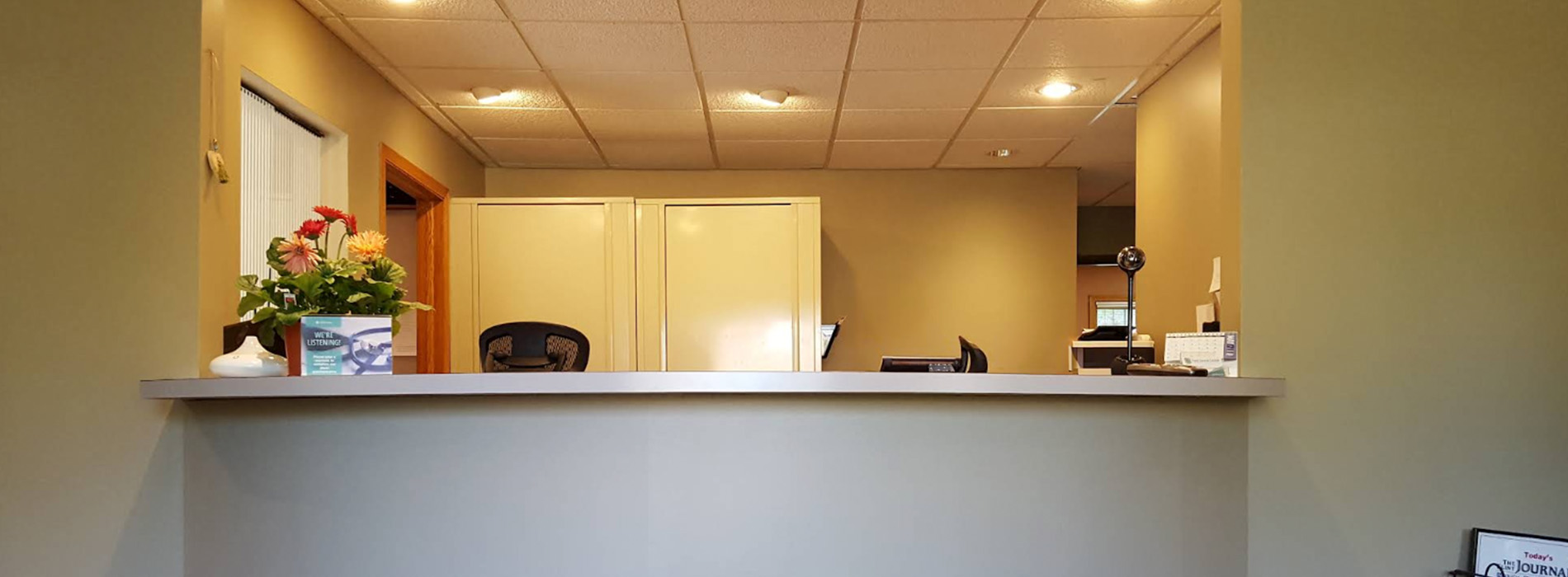 The image shows an interior view of a room with a desk and a reception area, featuring a countertop, chair, and a computer monitor.