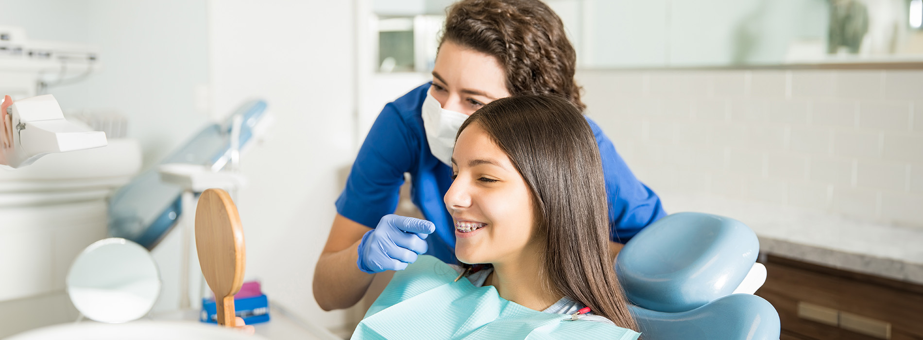 In the image, there are two individuals in a dental office setting a woman is seated in a dental chair with her head resting on a dental device, while another person, presumably a dentist or dental hygienist, stands nearby holding a mirror and smiling at the camera.