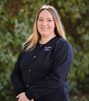 The image features a woman standing outdoors, wearing a black shirt with a name tag on her right side and a black apron, posing for the camera with a slight smile.