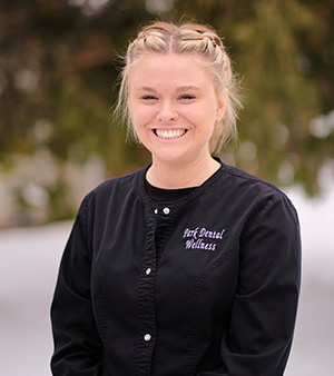 A young woman with blonde hair wearing a black shirt with a name tag and a stethoscope, smiling at the camera.
