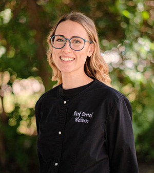 A woman wearing glasses stands smiling before a tree, dressed in a black shirt with white text and a name tag, posing for a portrait.