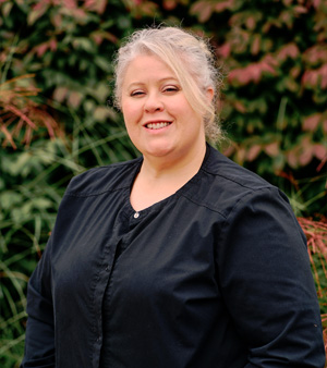 This is a portrait of a woman posing outdoors with a smile, standing against a backdrop featuring plants and foliage.