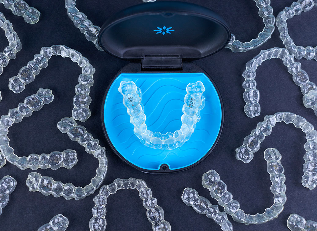 An up-close photograph showing a blue-lit mouthguard with clear plastic aligners next to it, set against a dark background with scattered gemstones.