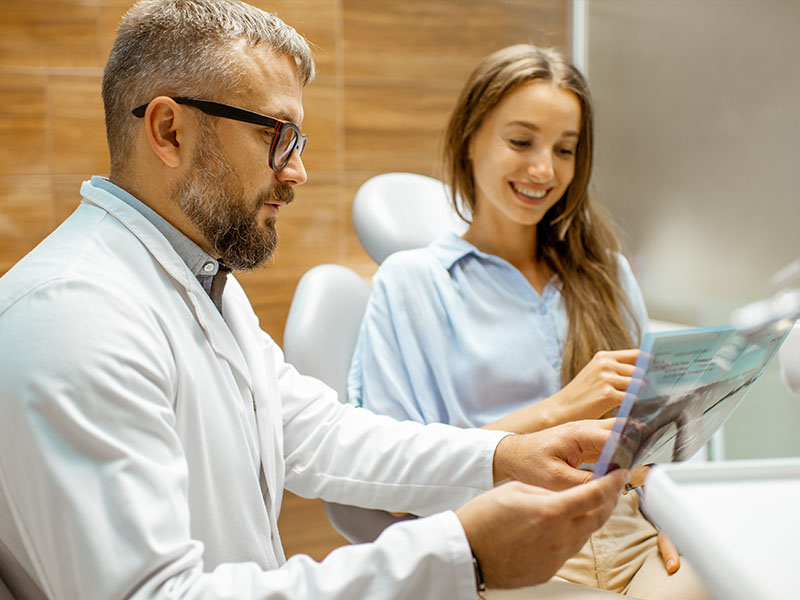 The image shows a dentist in his office with a patient, presenting an informative booklet about dental care.