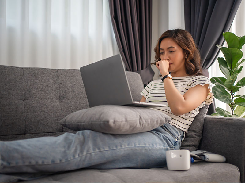 A young woman sitting on a couch with a laptop on her lap, looking down at the screen with a hand on her face, possibly in deep thought or concentration.