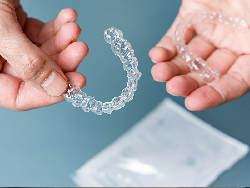 The image displays a transparent dental retainer being held by a human hand against a blue background.