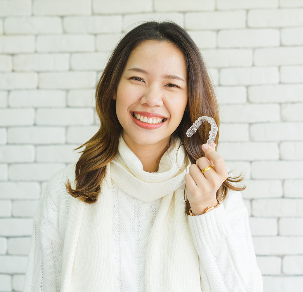 The image shows a smiling woman holding up a toothbrush with her right hand while standing in front of a brick wall background.