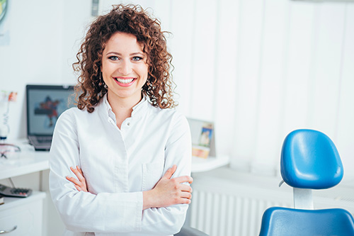 The image shows a smiling woman with curly hair standing in front of a dental office chair, wearing a white shirt and black pants, posing for a portrait with her arms crossed.