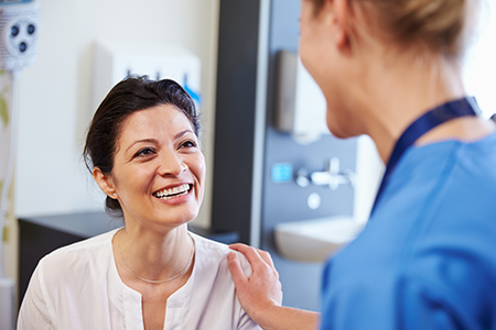 The image shows two people in a medical setting a woman with dark hair, wearing a white shirt and smiling broadly at the camera, stands next to another individual who appears to be a healthcare professional, possibly a nurse or doctor, based on their attire and the context of the scene.