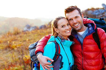 A man and woman outdoors, smiling and embracing each other, with the man wearing a backpack, both dressed for hiking or outdoor adventure.