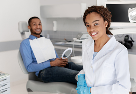 Woman in a dental office sitting on a chair with a man standing behind her, both wearing masks and smiling at the camera.