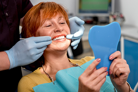 A woman sitting in a dental chair with a blue mouthguard, smiling at the camera while holding up a toothbrush with a toothpaste-like substance on it, with a dental hygienist in background preparing her teeth for cleaning.