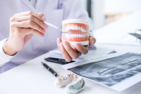The image shows a dentist holding up a model set of teeth with a magnifying glass, examining them closely against a backdrop of dental tools and equipment.