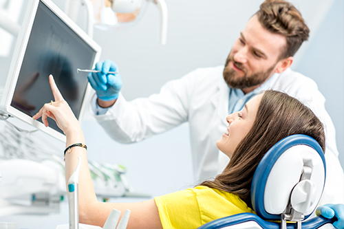 A person sitting in a dental chair with a dentist standing beside them, both wearing masks, in an office setting with medical equipment and screens visible.