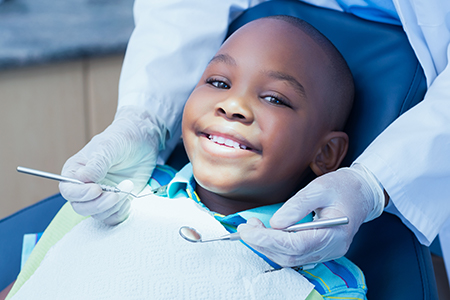 The image shows a young child sitting in a dental chair with a smiling expression, receiving dental treatment from a dentist wearing protective gloves and using dental instruments.