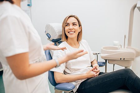 The image shows a woman sitting in a dental chair with a smile on her face, being attended to by a dental professional who appears to be a dentist, with other dental professionals in the background.
