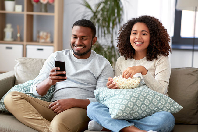 A man and woman sitting on a couch, smiling and enjoying popcorn together.